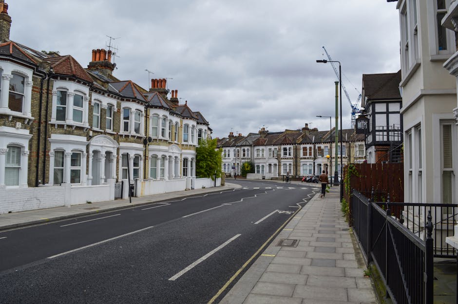 A quiet residential street scene during daytime showing terraced houses with Victorian-style architecture, including bay windows and decorative elements, on the left side of the image. The houses are painted in light colors with red tiled roofs and chimneys. On the right side, there are more modern buildings with dark timber framing and white facades. The pavement runs along the right side of the street, with a black metal railing and a few small plants visible. The street is empty except for a person walking in the distance. Several cars are parked along the curb on the far side of the street, and a tall streetlight is positioned near the center of the image, with a crane visible in the background. The sky is overcast with grey clouds. This scene captures a typical urban area suitable for a house removal or furniture transport process, with potential loading areas on the street or pavement close to the properties. Small construction or repair elements are visible, indicating ongoing development or maintenance, consistent with relocation logistics managed by Removal Van Hounslow.