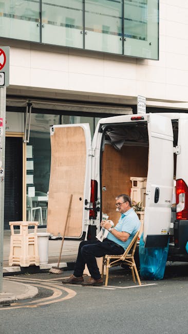 A man wearing glasses and a light blue shirt is seated on a wooden chair outside a white commercial van parked on the street in front of a modern building with large glass windows. The van's rear doors are open, revealing a partially loaded interior with wrapped packages, cardboard boxes, and furniture such as a small wooden table and a wooden plank leaning against the van. Inside the van, there are additional packing materials, including cardboard and protective wrapping. The scene depicts the process of home relocation or furniture transport, with the man taking a break or preparing items for loading or unloading. The environment shows a paved street with a yellow and black road marking nearby, and a signpost is visible above the van. The overall setting reflects a professional moving process supported by Removal Van Hounslow, aligning with house removals, packing, and transportation services.