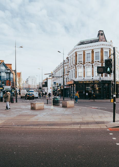 A wide view of Hounslow High Street during daytime, showcasing a busy urban street scene with pedestrian crossing signals, people walking on the sidewalks, and several buildings including a prominent three-story corner establishment with a sign reading 'The World's End.' The street features a mix of retail shops, some with visible signage, and a vintage-style double-decker bus is parked on the left side. In the foreground, the pavement is paved with bricks, with a few large wooden planters positioned near the crossing. A white van from Removal Van Hounslow is visible in the background, likely involved in home relocation or furniture transport activities as part of a house moving process. The scene is illuminated by diffused daylight through partly cloudy skies, with streetlights and overhead tram wires visible. The environment captures the typical busy atmosphere of house removals in the area, with a focus on transportation, urban logistics, and moving-related infrastructure.
