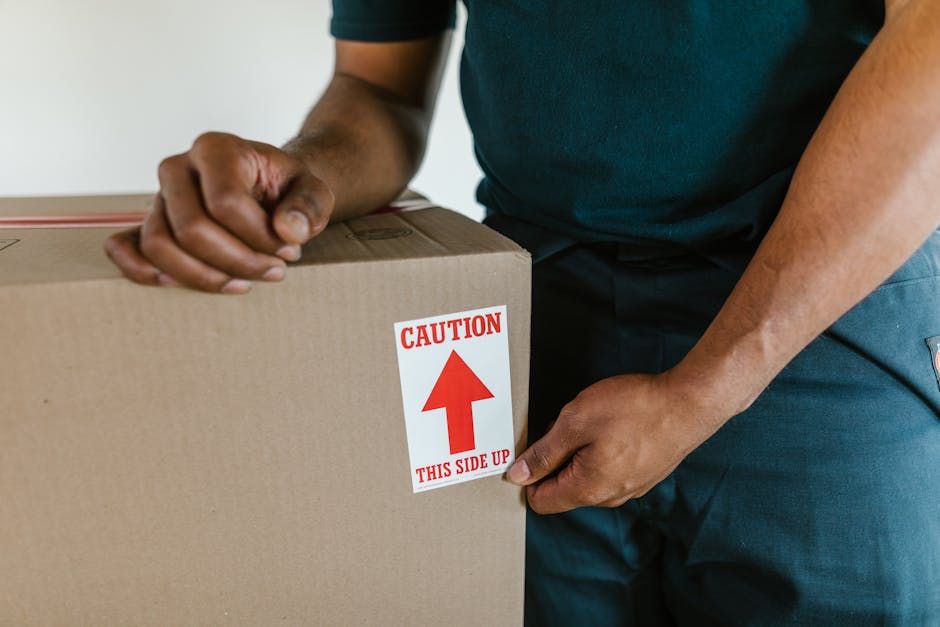Close-up of a person wearing a dark teal shirt and dark pants, carefully holding a large cardboard box with a white label that has red text reading 'CAUTION THIS SIDE UP' and an upward-pointing arrow. The individual appears to be in the process of packing or preparing the box for a house removal, with their hands gripping the sides of the box. The background suggests an indoor environment, possibly a home or storage area, with a focus on the correct handling of packing materials during furniture transport or moving logistics. The scene illustrates the importance of proper packing and handling in professional removals, as carried out by companies like Removal Van Hounslow, to ensure safe and efficient furniture transport during home relocation or packing and moving services.