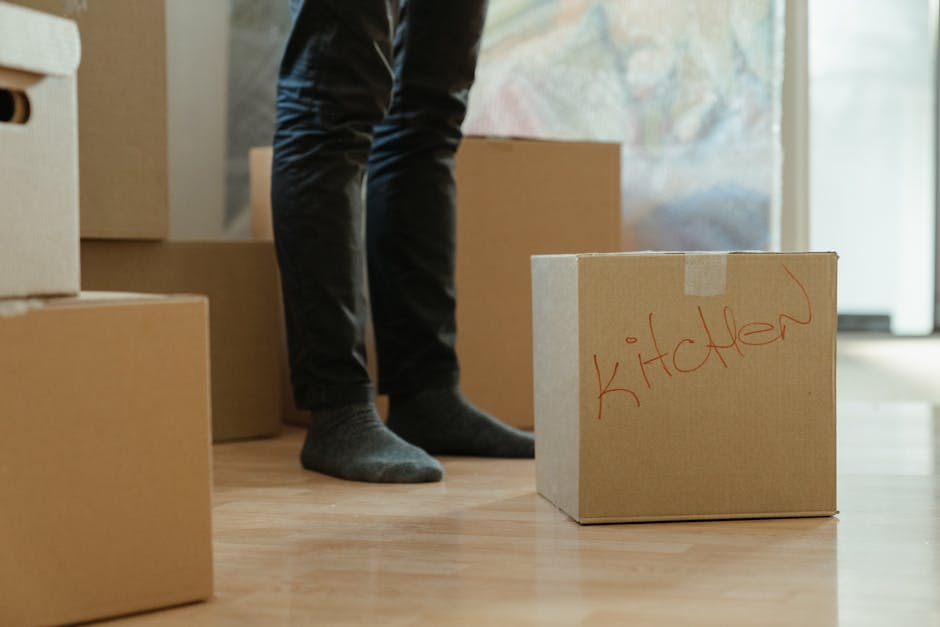 Close-up of a person wearing a dark teal shirt and dark pants, carefully holding a large cardboard box with a white label that has red text reading 'CAUTION THIS SIDE UP' and an upward-pointing arrow. The individual appears to be in the process of packing or preparing the box for a house removal, with their hands gripping the sides of the box. The background suggests an indoor environment, possibly a home or storage area, with a focus on the correct handling of packing materials during furniture transport or moving logistics. The scene illustrates the importance of proper packing and handling in professional removals, as carried out by companies like Removal Van Hounslow, to ensure safe and efficient furniture transport during home relocation or packing and moving services.
