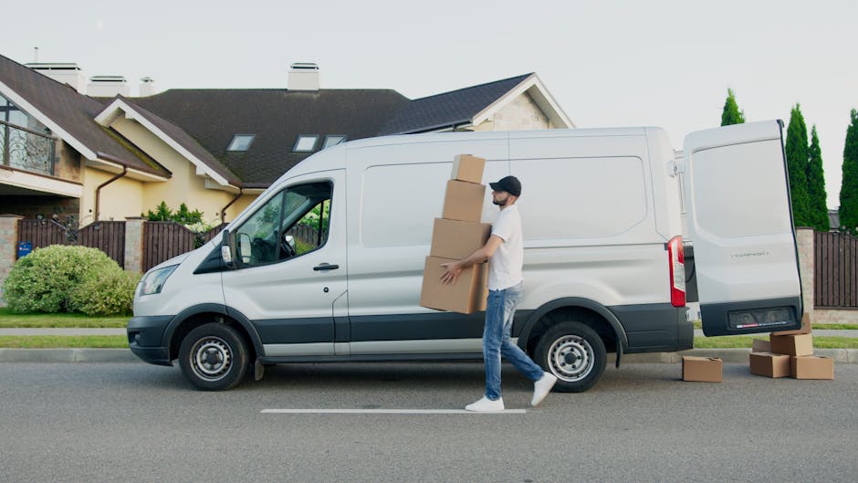 A man wearing casual clothing and a baseball cap is loading three large cardboard boxes into the side door of a white commercial van parked on a residential street. Additional boxes are placed on the