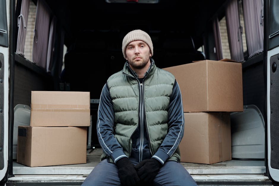 A man wearing a beige knit beanie, a green and navy blue puffer jacket, and black gloves is seated inside the open rear of a moving van, which is parked on a pavement. The van's interior contains several cardboard boxes, some stacked and others placed individually, ready for a home relocation. The boxes vary in size and are sealed with clear packing tape. The loading area behind the man shows parts of the van's dark interior, with a glimpse of the vehicle's sliding doors open to the outside. The scene suggests a furniture transport process as part of packing and moving activities handled by Removal Van Hounslow, highlighting the loading phase of house removals and professional moving logistics.