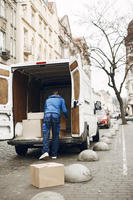 A man wearing a beige knit beanie, a green and navy blue puffer jacket, and black gloves is seated inside the open rear of a moving van, which is parked on a pavement. The van's interior contains several cardboard boxes, some stacked and others placed individually, ready for a home relocation. The boxes vary in size and are sealed with clear packing tape. The loading area behind the man shows parts of the van's dark interior, with a glimpse of the vehicle's sliding doors open to the outside. The scene suggests a furniture transport process as part of packing and moving activities handled by Removal Van Hounslow, highlighting the loading phase of house removals and professional moving logistics.